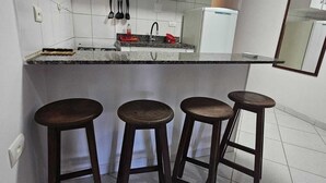 A compact kitchen with white cabinets, a granite countertop, a sink, and three wooden stools. - Suites Pontal dos Golfinhos (Ipojuca)