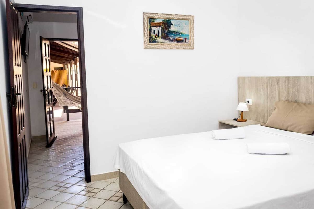 Une chambre d’hôtel avec un lit, une table de chevet avec une lampe, un tableau au mur et une vue sur la plage, visible par une porte ouverte.