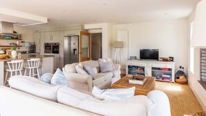 Ein modernes Wohnzimmer mit einer Couch, einem Couchtisch und einem Fernseher. Im Hintergrund befindet sich eine Küchenzeile mit Schränken und Geräten.
