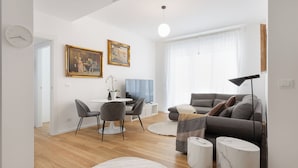 A modern living room with a grey sofa, a white coffee table, and a dining area with a white table and chairs. There are two paintings on the wall, a ceiling light, and a floor lamp.