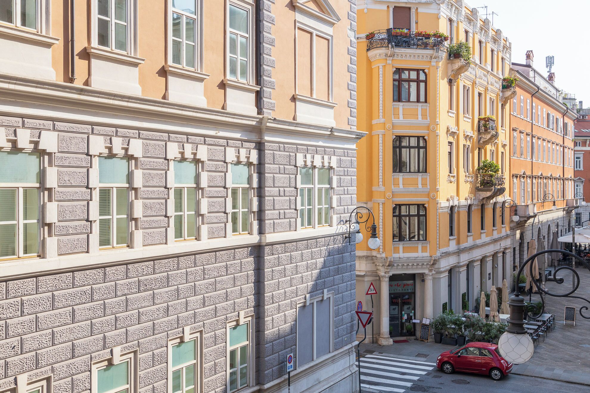 Straßenansicht mit einem roten Auto, das am Straßenrand geparkt ist, Gebäuden im klassizistischen Stil und einer Fußgängerüberweg.