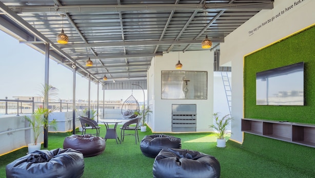Une terrasse sur le toit, avec un revêtement de gazon synthétique, des poufs noirs, une table avec des chaises, des plantes en pot et un téléviseur fixé sur un mur vert.