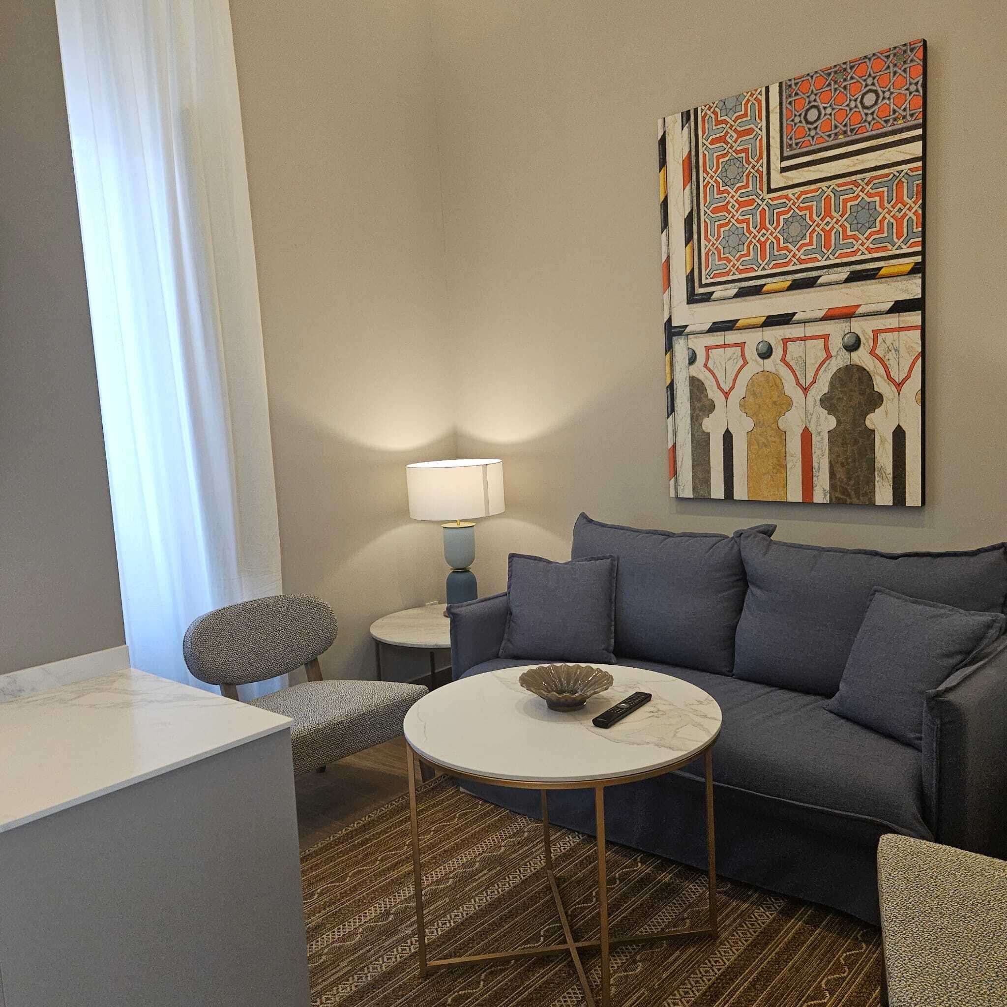 Una sala de estar moderna con un sofá gris, una mesa redonda para café y un tapete estampado. Hay una lámpara sobre una mesita auxiliar y una pieza decorativa en la pared sobre el sofá.