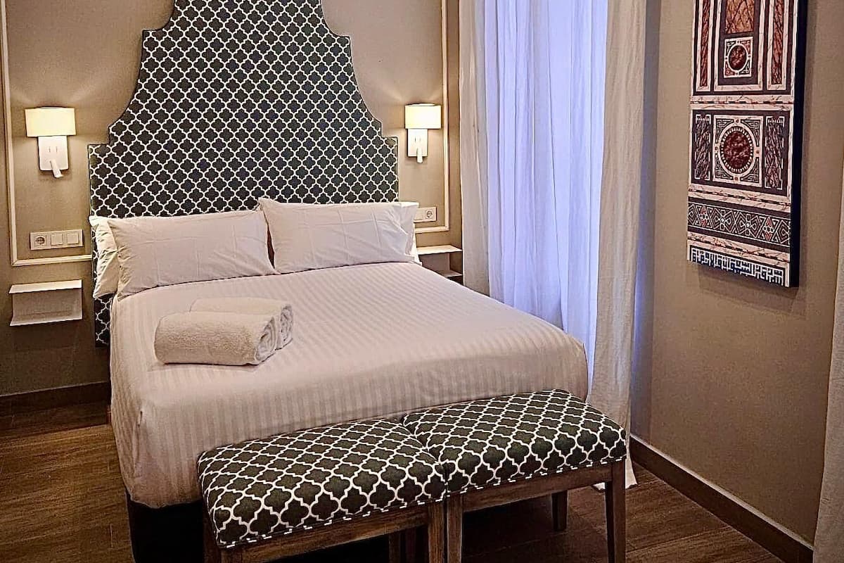 Een keurig opgeruimde hotelkamer met een groot bed, nachtlampjes, een bankje en decoratieve wandpanelen.