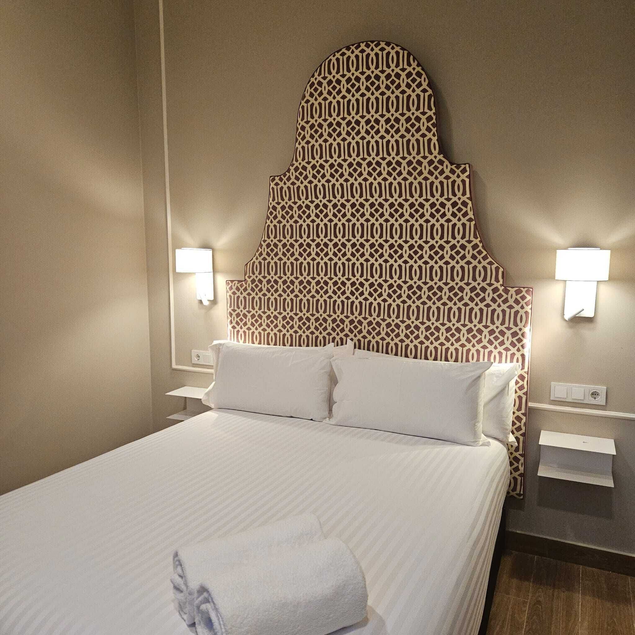 Habitación de hotel con un cabecero estampado, dos lámparas de noche, una cama bien hecha con ropa blanca y una toalla sobre la cama.