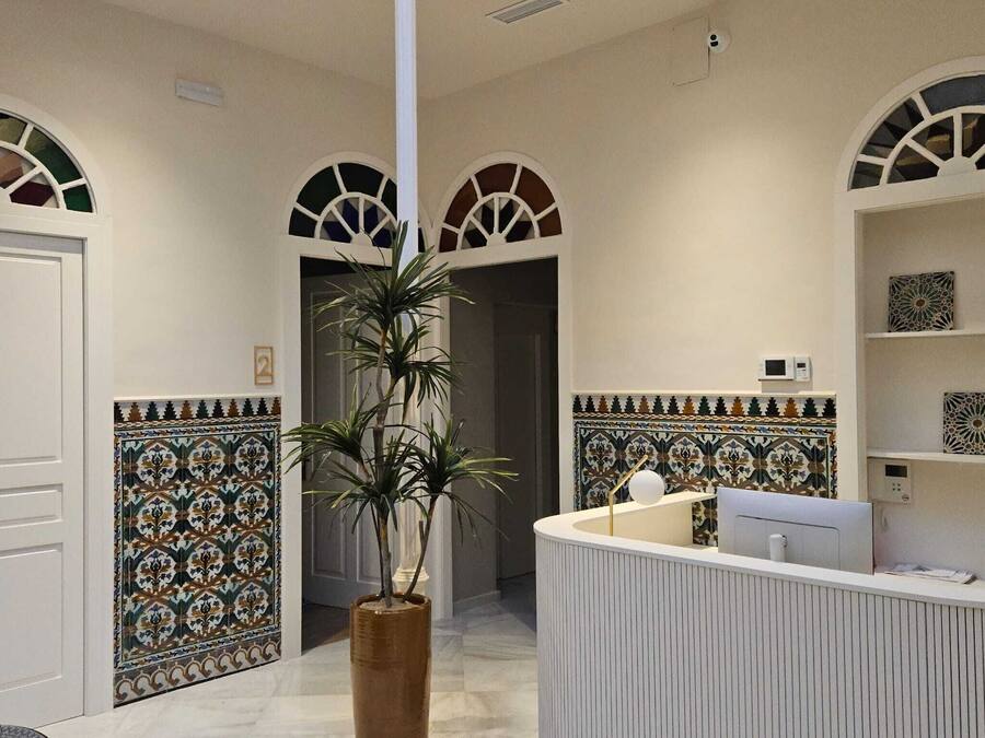 Un área de recepción con un mostrador blanco, una planta grande en maceta y paredes decoradas con azulejos.