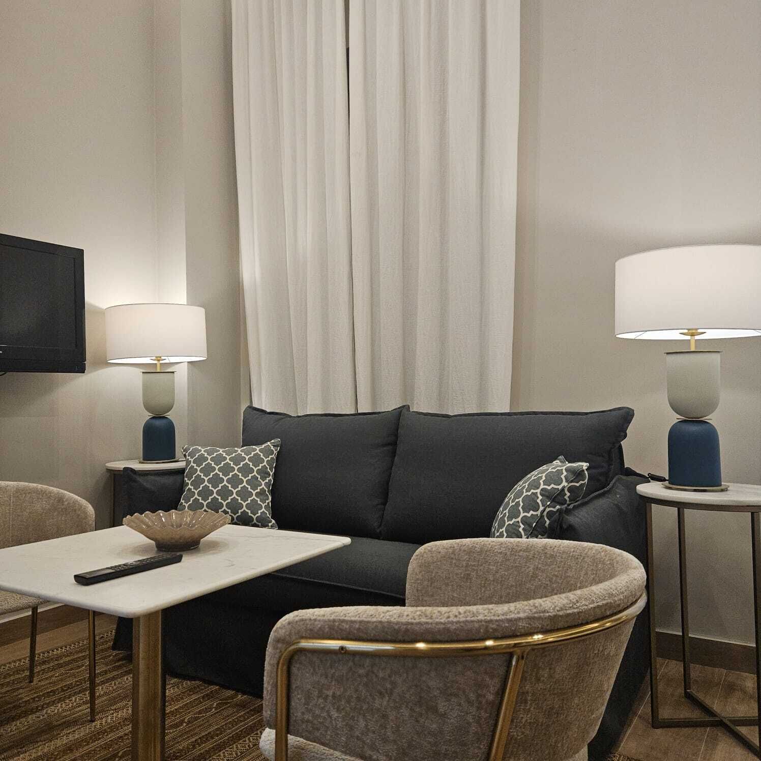 Una habitación de hotel moderna con un sofá gris oscuro, dos sillones, una mesita blanca y dos lámparas de noche.