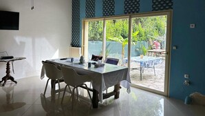 A dining area with a table and chairs, a small side table, and a large sliding glass door leading to an outdoor patio. - Villa F3 entre mer et Montpellier (Lattes)