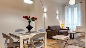 A modern living room with a dining area, featuring a table with chairs, a sofa, a lamp, and a vase with red flowers.