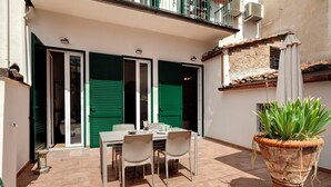 An outdoor patio with a table and chairs, a potted plant, and a building entrance with green shutters.