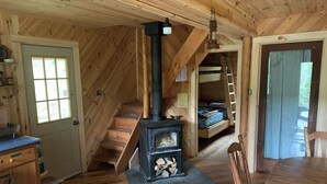 L'intérieur d'une cabane en bois est aménagé avec un poêle à bois central, une table à manger et un escalier menant à une chambre à coucher située en mezzanine.