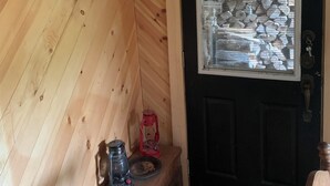 Un intérieur en bois avec une porte noire, une fenêtre donnant sur une pile de bûches et une petite table sur laquelle se trouvent une lanterne et une bouilloire.