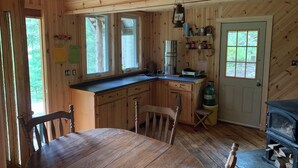 Une cabane en bois comprenant une table à manger, des chaises, un coin cuisine et un poêle à bois.
