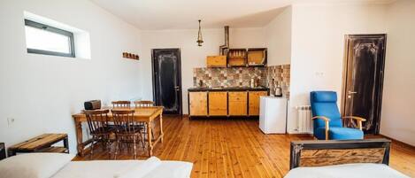 Una habitación compacta con suelo de madera, un comedor con sillas, una cama con cabecero, una kitchenette con armarios y una silla azul.