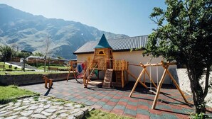 A playground with wooden structures, a stone pathway, and a building with a tiled roof. - Green Sheep Boutique Hotel (Stepantsminda)