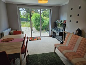 A living room with a piano, a sofa, a dining table with chairs, and a view through a glass door to a garden. - Charming family home in the heart of Sanderstead. (Greater London)