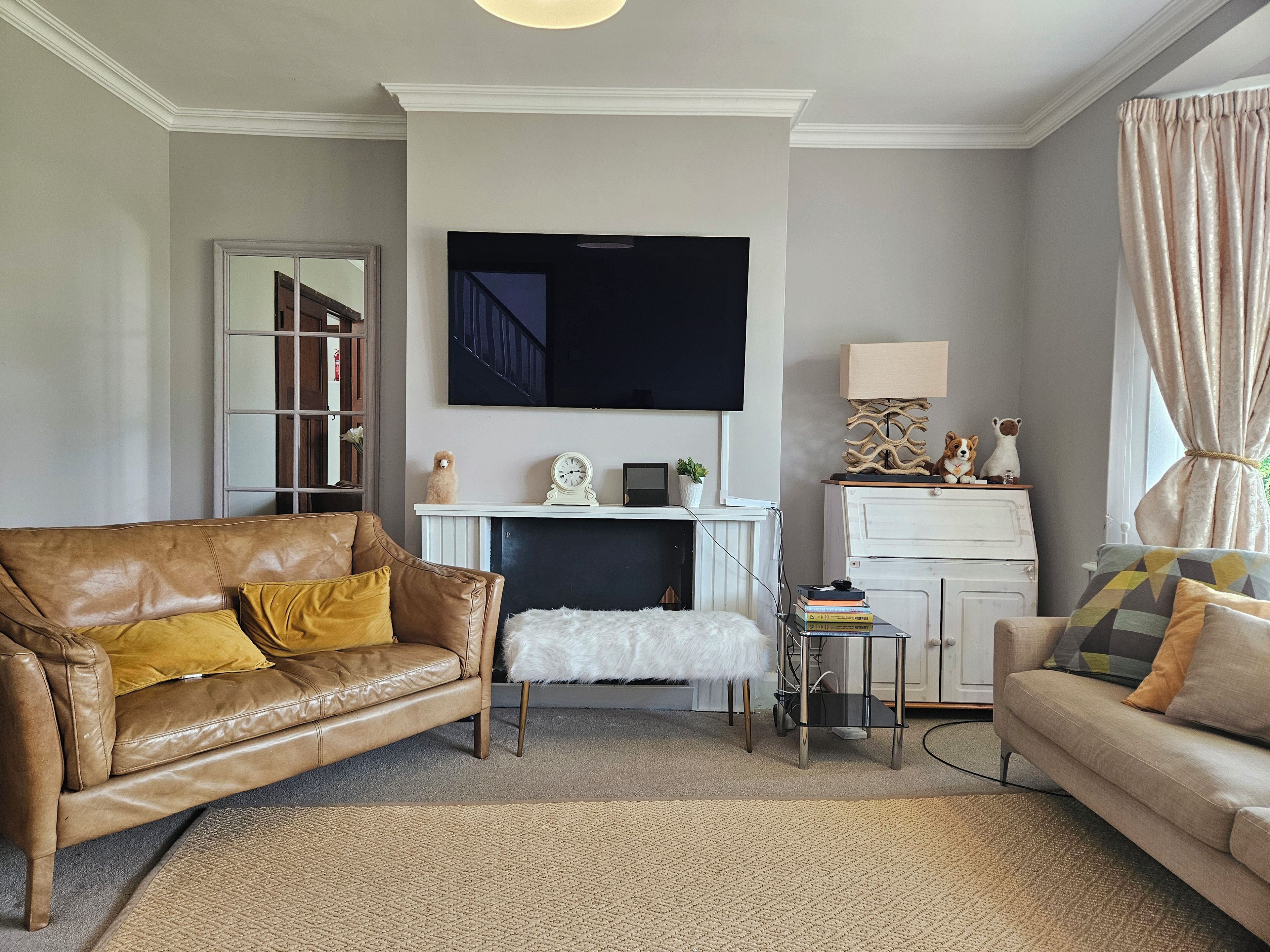 Ein Wohnzimmer mit einer braunen Ledersofa, einem weißen, flauschigen Hocker, einem beigen Teppich, einem an der Wand montierten Fernseher, einem kleinen Tisch mit Dekoration und einem weißen Schrank mit einer Lampe.