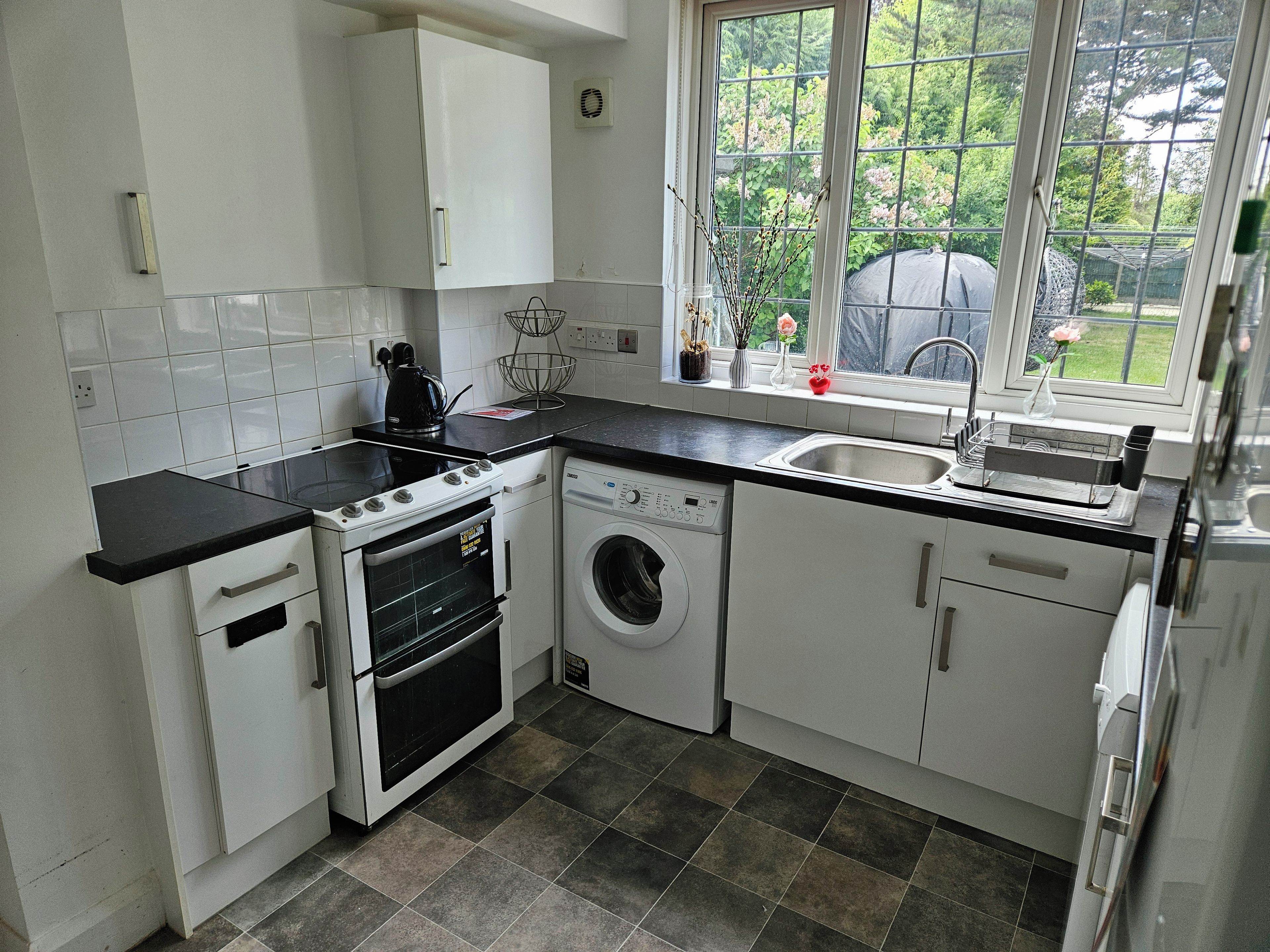 A compact kitchen with white cabinets, a stove, a washing machine, and a window with a view of greenery.
