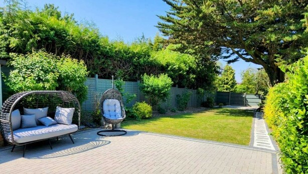 A patio with a wicker sofa and a hanging chair, surrounded by lush greenery and a paved walkway. - Charming family home in the heart of Sanderstead. (Greater London)