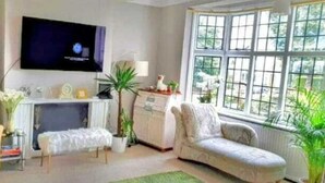 A living room with a television mounted on the wall, a sofa, a small table, and a window with a view of greenery. - Charming family home in the heart of Sanderstead. (Greater London)