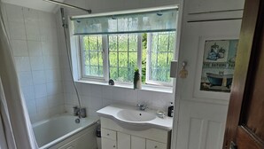A bathroom with a bathtub, a sink, and a window with a view of greenery. - Charming family home in the heart of Sanderstead. (Greater London)
