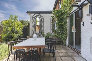 Une terrasse avec une table et des chaises, un mur blanc avec un portail en métal noir et un mur de briques orné de verdure.