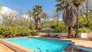 A rectangular swimming pool with a tiled edge, surrounded by a tiled deck. There are lounge chairs and a shaded area with a white umbrella. - Villa Falcone (Piazza Armerina)