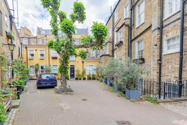 A narrow street with a parked car, brick buildings, and a tree with hanging plants.