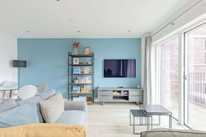 A modern living room with a sofa, TV, bookshelf, and a balcony with a view. - Modern Canal-Side 1BD Flat, Heart of Hackney (Greater London)