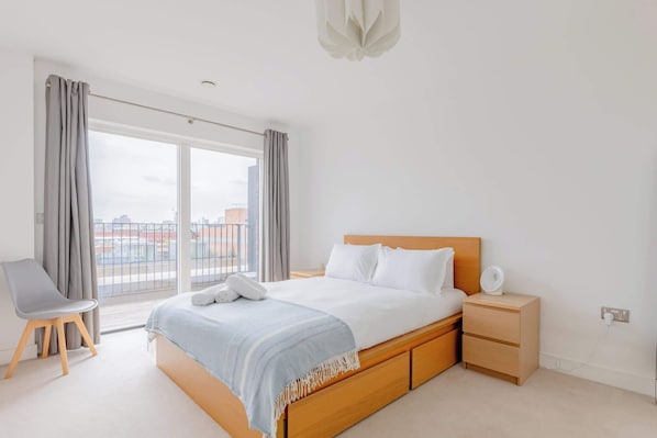 A bedroom with a large bed, a bedside table, a chair, and a view of the city through the window.