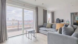 A modern living room with a grey sofa, a small table, and a balcony with a city view. - Modern Canal-Side 1BD Flat, Heart of Hackney (Greater London)