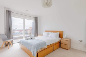 A bedroom with a large bed, a bedside table, a chair, and a view of the city through the window.