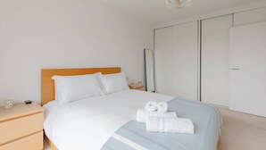 A neatly made bed with white linens and two rolled towels, a wooden headboard, a bedside table with a lamp, and a wardrobe with sliding doors.