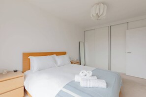 Un lit bien fait, avec du linge de lit blanc et deux serviettes enroulées, une tête de lit en bois, une table de chevet avec une lampe et une armoire aux portes coulissantes.
