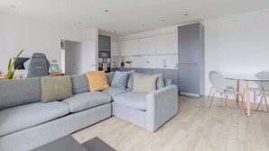 A modern living room with a grey sectional sofa, a glass dining table with chairs, and a kitchen area in the background.