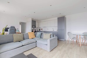 A modern living room with a grey sectional sofa, a glass dining table with chairs, and a kitchen area in the background. - Modern Canal-Side 1BD Flat, Heart of Hackney (Greater London)