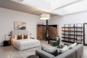 A modern bedroom with a large bed, a green armchair, a grey sofa, a fireplace, and a bookshelf.