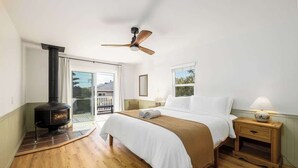 A bedroom with a bed, a nightstand, a wooden floor, a ceiling fan, a window, and a fireplace. - Carlsbad Cottage - Remodeled (Carlsbad)