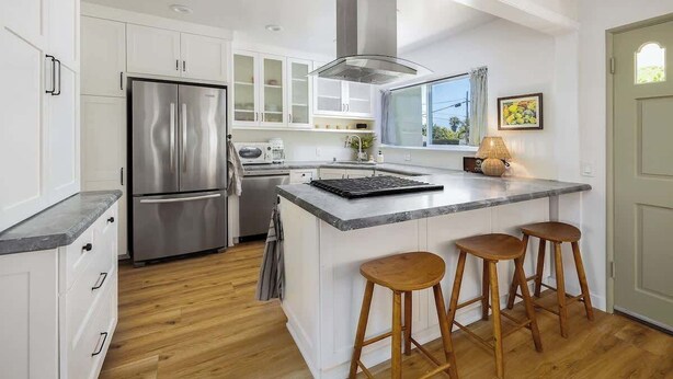 A modern kitchen with white cabinetry, a stainless steel refrigerator, a large island with stools, and a window providing natural light. - Carlsbad Cottage - Remodeled (Carlsbad)