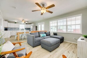 A modern living room with a grey sectional sofa, wooden chairs, a ceiling fan, and a kitchen in the background. - The Mermaid Cove Ocean View Beach House Surfside (Freeport)