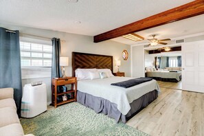 A bedroom with a large bed, a wooden headboard, a nightstand, a lamp, a mirror, and a ceiling fan. - The Mermaid Cove Ocean View Beach House Surfside (Freeport)