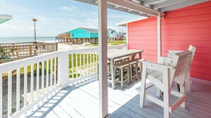A wooden deck with a white railing, a red building, and a turquoise house with a pier. - The Mermaid Cove Beach House with Ocean Views (Freeport)