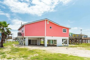 A red building with a white base, elevated on stilts, with a covered parking area and a small deck. - The Mermaid Cove Ocean View Beach House Surfside (Freeport)