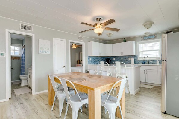 A modern kitchen with white cabinets, a wooden dining table with white chairs, a ceiling fan, and a window with a view. - The Mermaid Cove Beach House with Ocean Views (Freeport)