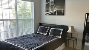 Une chambre à coucher avec un lit, un ventilateur de plafond, une fenêtre avec des stores, une lampe sur une table de chevet et un tableau encadré au mur.