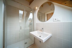 Un baño con ducha de vidrio, un lavamanos blanco y un espejo redondo.