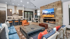 A modern living room with a stone fireplace, comfortable seating, and a kitchen in the background. - Vail Chalet | Platinum-Rated 4BR Luxury Home, Cascade (Vail)