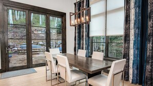 A dining area with a wooden table and white chairs, large windows with curtains, and a view of a forested outdoor area. - Vail Chalet | Platinum-Rated 4BR Luxury Home, Cascade (Vail)