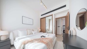 A modern hotel room with a large bed, a bedside table, a dresser, a mirror, and a view of the bathroom and kitchen through an open door. - StayShort - Luxury 1-Bedroom JVC Apartment with Balcony & Pool (JVC)