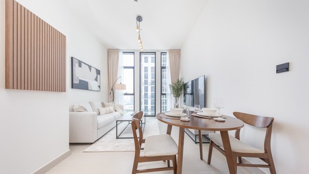 A modern living room with a sofa, a dining table with chairs, a TV, and a large window with curtains. - StayShort - Luxury 1-Bedroom JVC Apartment with Balcony & Pool (JVC)
